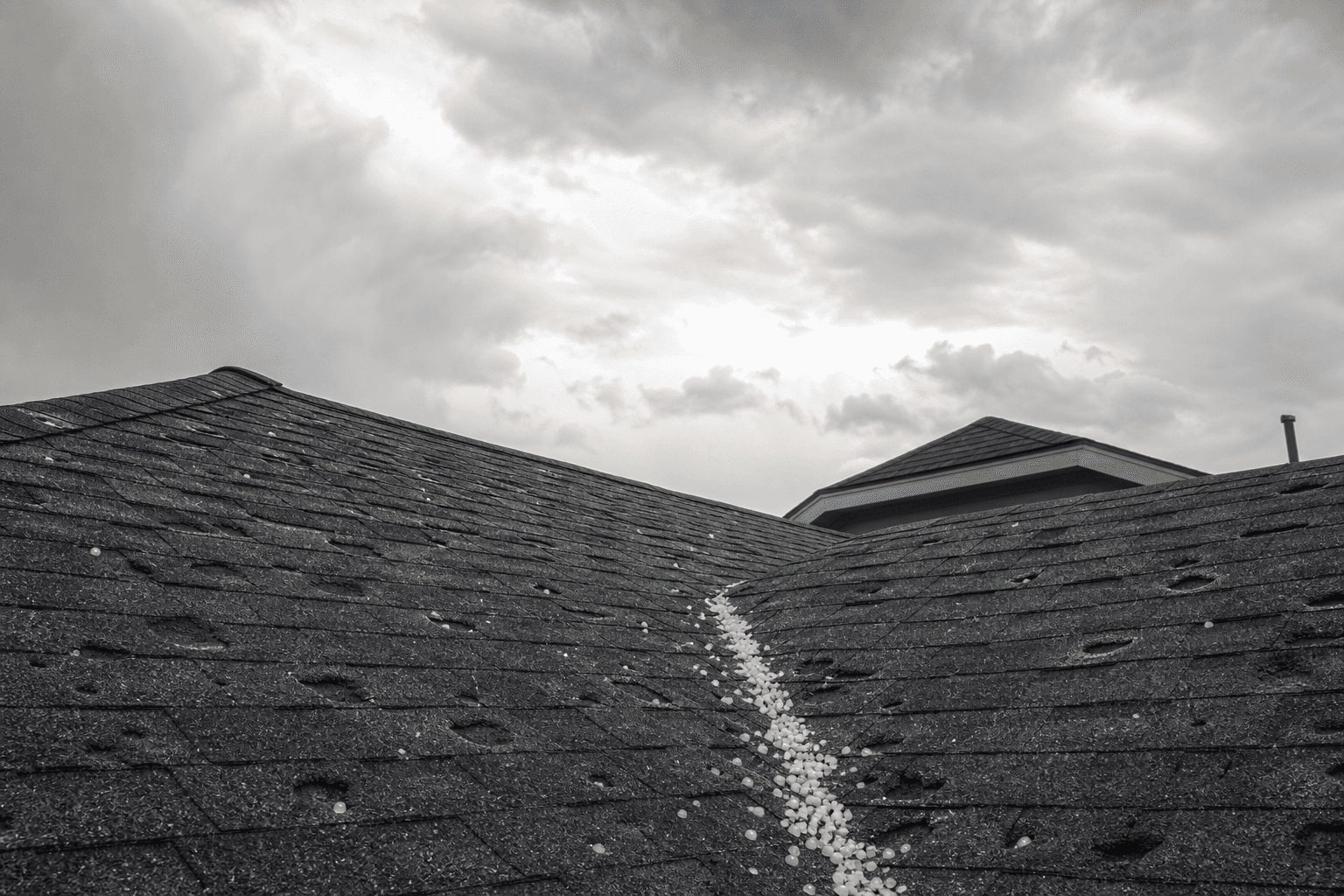 Hail stones caught in the valley of a dented shingle roof under a Kansas City storm sky